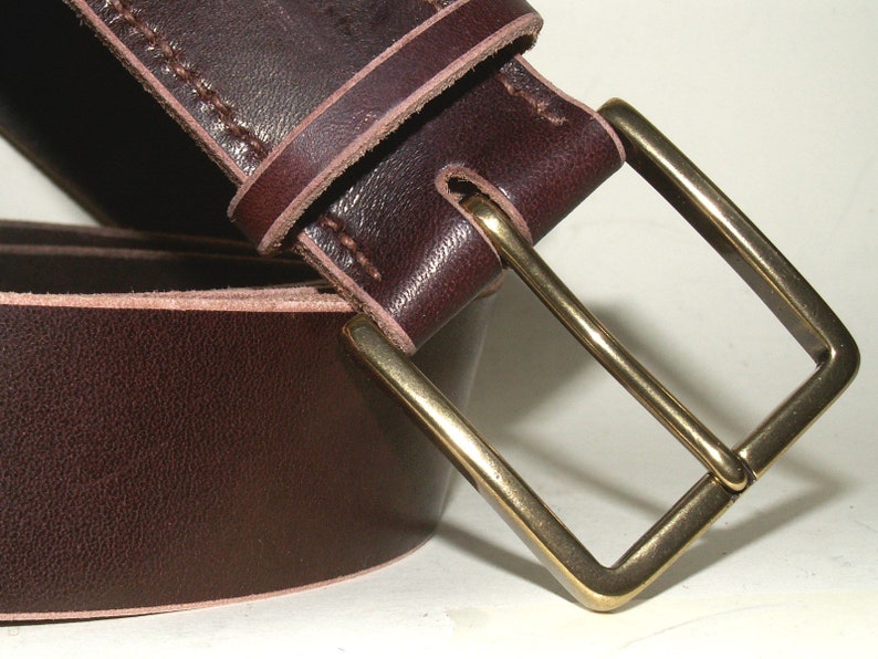 May include: Close-up of a dark brown leather belt with a rectangular brass buckle. The belt has visible stitching along the edges and a smooth, polished surface. The buckle is a classic, simple design.