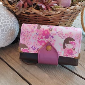 May include: A pink and brown wallet with a floral pattern and a pink leather flap. The wallet has gold accents and a small metal button.
