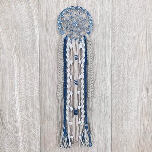 Könnte beinhalten: Ein blauer und weißer Traumfänger mit einem kreisförmigen Design und mehreren Strängen aus weißem und beigefarbenem Garn, die von unten hängen. Der Traumfänger hängt vor einem hellen Holzhintergrund.