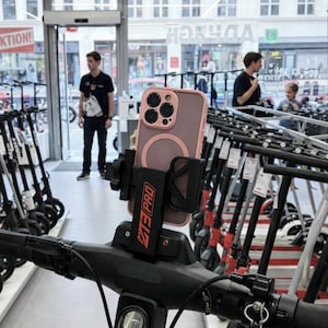 May include: A black scooter handlebar with a phone holder and a phone in a pink case. The phone holder has the text "MT3 PRO" in red. The background shows a store with scooters and people.