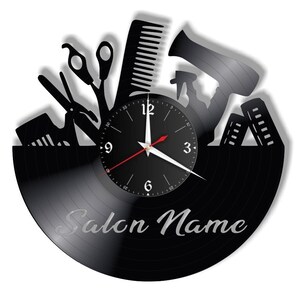 Puede incluir: Reloj de vinilo negro con temática de salón. El reloj presenta un peine, tijeras, un secador de pelo y una botella de laca para el cabello. La esfera del reloj es negra con números y manecillas blancas. El texto "Salon Name" está impreso debajo de la esfera del reloj.