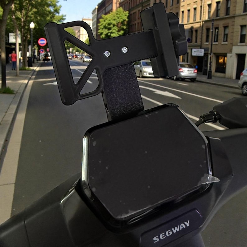 May include: Black Segway scooter with a phone mount attached to the handlebars. The mount is made of black plastic and has a textured grip. The scooter is parked on a city street with cars and buildings in the background.