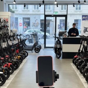 May include: An interior shot of an e-scooter store. The foreground shows the handlebars of a black scooter with a phone mount holding a pink-edged phone. Rows of scooters in various colors line the store, with a person at the counter in the background.