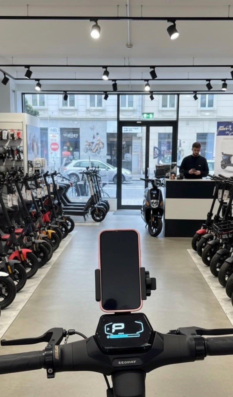 May include: An interior shot of an e-scooter store. The foreground shows the handlebars of a black scooter with a phone mount holding a pink-edged phone. Rows of scooters in various colors line the store, with a person at the counter in the background.