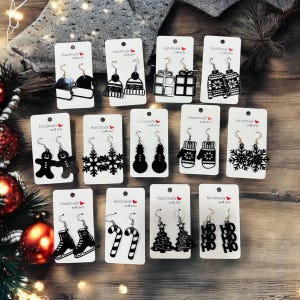 Puede incluir: Doce pares de pendientes negros con temática navideña en tarjetas blancas con el texto "Handmade with love". Los pendientes presentan diseños como un muñeco de nieve, un copo de nieve, un árbol de Navidad, un bastón de caramelo, una media, una manopla, una caja de regalo, un hombre de jengibre y las palabras "HO HO".