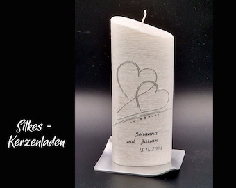 May include: A white pillar candle with a textured surface, featuring two black line-art hearts and the text "Johanna und Julian 13.11.2021" printed on the front. The candle sits on a square silver base and has a single white wick at the top.