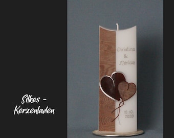 May include: A white pillar candle with a concave top features a brown wood-grain panel, two overlapping heart decorations, and the text Christina & Markus with the date 10.10.2020. The candle stands on a circular silver base.