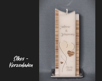 May include: A white wedding candle with a wooden trim and two silver hearts. The candle is inscribed with the names "Sabine & Johannes" and the date "25.05.2021".