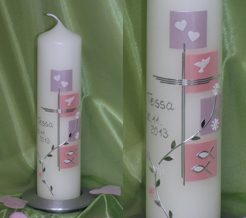May include: White baptism candle with a silver cross design. The candle has pink and purple squares with silver doves, hearts, and fish. The text "Tessa 09.11. 2013" is written in silver.