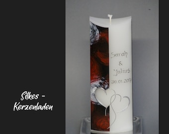 May include: White pillar candle with a red and black abstract design on one side. The other side has the text "Sarah & Julius 30.01.2017" and two silver hearts. The candle is on a silver base.