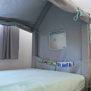 K&ouml;nnte beinhalten: Ein graues und wei&szlig;es Zelt-&auml;hnliches Baldachin-Bettgestell mit einer hellblauen Fleece-Decke und einem kleinen Fenster mit einem Sternendesign. Das Bett hat eine Tasche an der Seite mit B&uuml;chern, darunter eines mit dem Titel "Elmer".