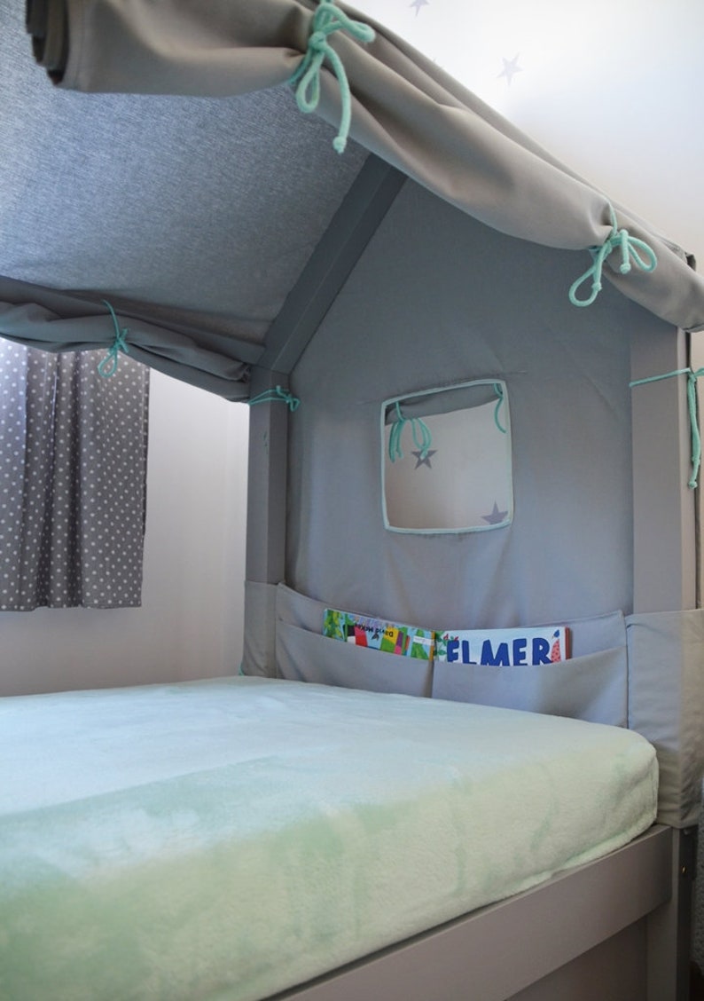 K&ouml;nnte beinhalten: Ein graues und wei&szlig;es Zelt-&auml;hnliches Baldachin-Bettgestell mit einer hellblauen Fleece-Decke und einem kleinen Fenster mit einem Sternendesign. Das Bett hat eine Tasche an der Seite mit B&uuml;chern, darunter eines mit dem Titel "Elmer".