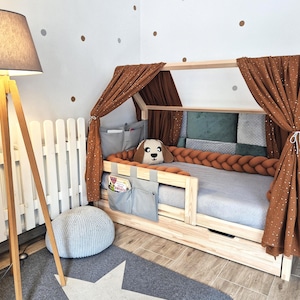 Op de afbeelding: Een houten bed met een luifel, met bruine gordijnen met witte sterrenpatronen. Het bed heeft een grijze matras, een gevlochten oranje bumper en een hondvormig kussen. Een grijs tapijt met een witte ster ligt ervoor.