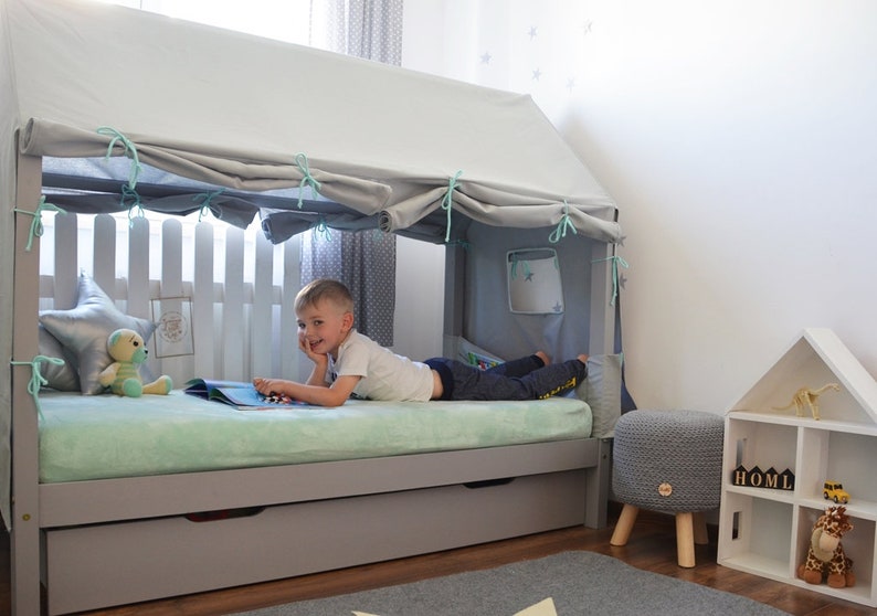 K&ouml;nnte beinhalten: Ein graues und wei&szlig;es Holzbettgestell mit einem Baldachin und einer hellgr&uuml;nen Matratze. Ein Kind liegt auf dem Bett und liest ein Buch. Es gibt eine graue und wei&szlig;e Aufbewahrungspolster und ein wei&szlig;es B&uuml;cherregal mit dem Wort "HOME" in Holzbl&ouml;cken.
