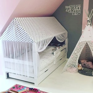 May include: A white bed frame with a canopy tent featuring a black and white star pattern. The bed has a white mattress and a white and gray patterned pillow. The bed is in a room with a pink wall and a gray wall with a wooden sign that says "Regarde UNE FOIS TOI & MOI Un JOUR! Celia". There is a white teepee tent with a white lace trim in the corner of the room.