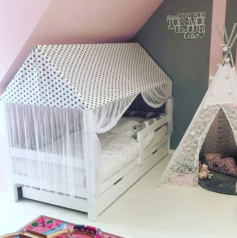 May include: A white bed frame with a canopy tent featuring a black and white star pattern. The bed has a white mattress and a white and gray patterned pillow. The bed is in a room with a pink wall and a gray wall with a wooden sign that says "Regarde UNE FOIS TOI & MOI Un JOUR! Celia". There is a white teepee tent with a white lace trim in the corner of the room.