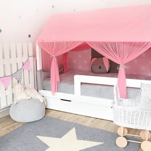 Puede incluir: Una cama de madera blanca con dosel rosa y un cochecito de mu&ntilde;ecas de mimbre blanco. La cama tiene un colch&oacute;n estampado en gris y blanco y una almohada estampada en gris y blanco. La cama est&aacute; decorada con una guirnalda rosa y gris y una alfombra de estrella blanca.