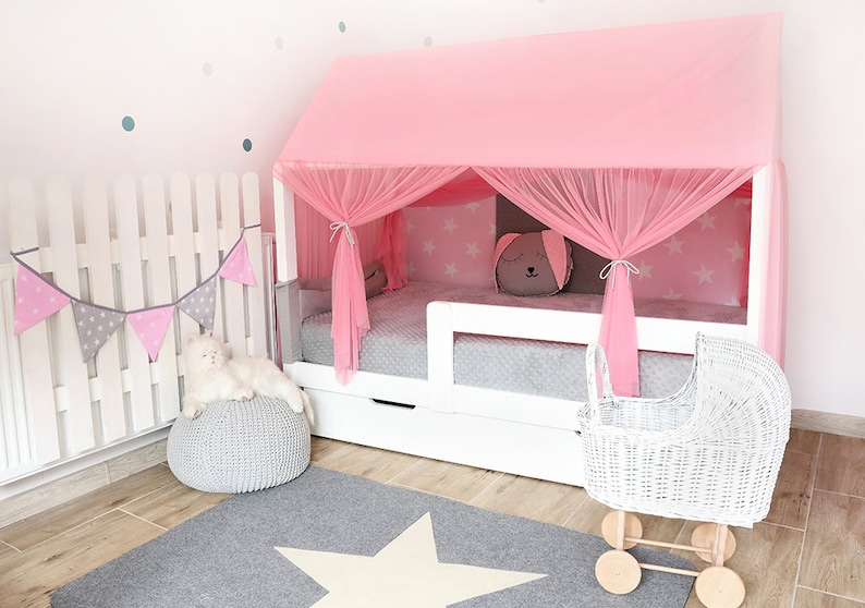 Puede incluir: Una cama de madera blanca con dosel rosa y un cochecito de mu&ntilde;ecas de mimbre blanco. La cama tiene un colch&oacute;n estampado en gris y blanco y una almohada estampada en gris y blanco. La cama est&aacute; decorada con una guirnalda rosa y gris y una alfombra de estrella blanca.