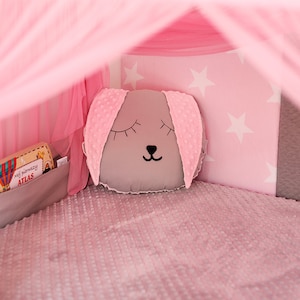 Puede incluir: Una tienda de juegos rosa y gris con una almohada gris con una linda cara de animal. La tienda tiene un patr&oacute;n de estrellas blancas en la pared trasera y un libro en un bolsillo en el lateral.