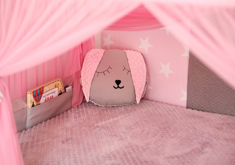 Puede incluir: Una tienda de juegos rosa y gris con una almohada gris con una linda cara de animal. La tienda tiene un patr&oacute;n de estrellas blancas en la pared trasera y un libro en un bolsillo en el lateral.