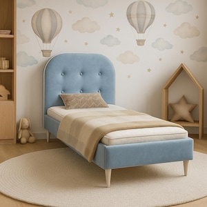 Pode incluir: Uma cama de solteiro estofada em azul claro com cabeceira com botões e um cobertor listrado bege e branco. A cama está em um quarto infantil com um tapete redondo e um mural de balões de ar quente.