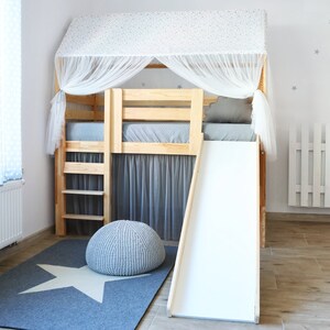 May include: A wooden loft bed with a white canopy and a white slide. The bed has a gray mattress and a gray rug with a white star on it. There is a gray pouf in front of the bed.