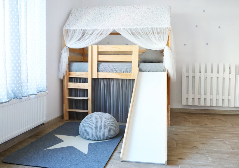 May include: A wooden loft bed with a white canopy and a white slide. The bed has a gray mattress and a gray rug with a white star on it. There is a gray pouf in front of the bed.