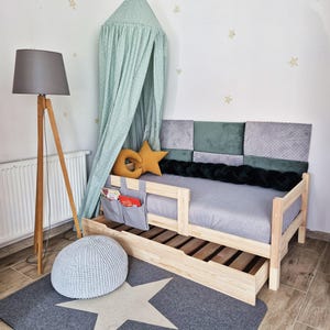 Könnte beinhalten: Ein Holzbettgestell mit einer grauen Matratze und einem grünen Baldachin. Das Bett hat eine Schublade und einen grauen Strickhocker. Das Bett ist mit Kissen in verschiedenen Grüntönen und Grau sowie einem gelben sternförmigen Kissen dekoriert.