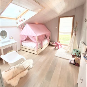 May include: A pink canopy bed with a white mattress and a white wooden frame. The bed is in a room with white walls and a wooden floor. There is a white rocking chair in the foreground and a white vanity with a mirror in the background.