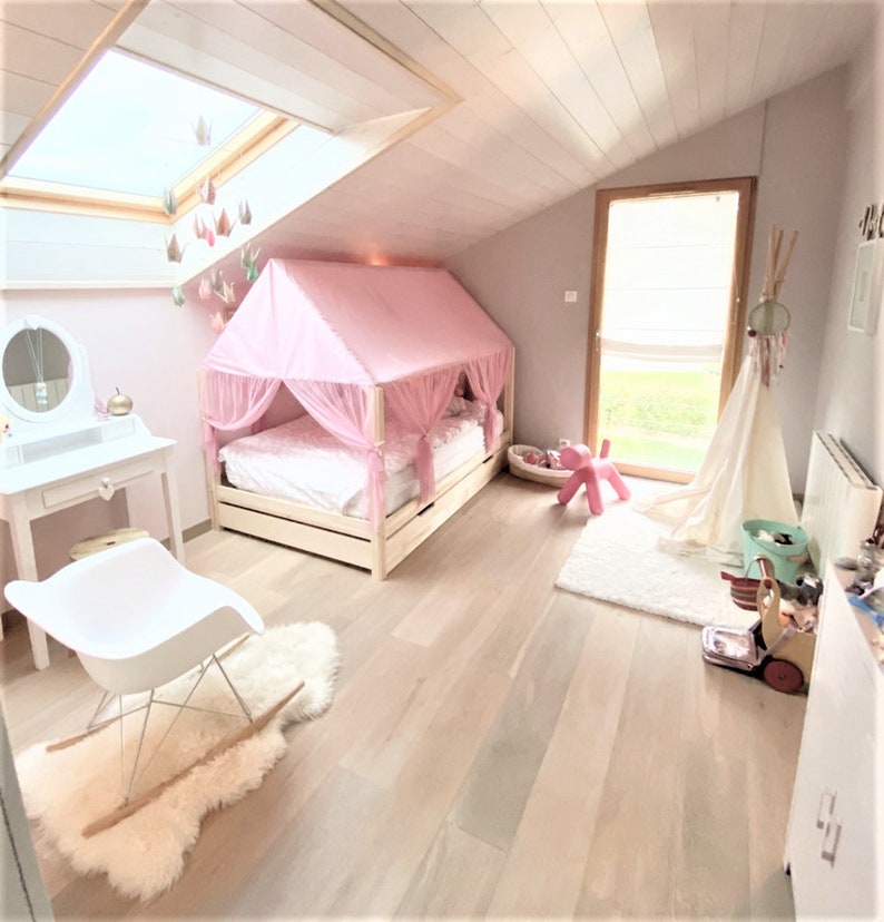 May include: A pink canopy bed with a white mattress and a white wooden frame. The bed is in a room with white walls and a wooden floor. There is a white rocking chair in the foreground and a white vanity with a mirror in the background.