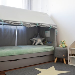 K&ouml;nnte beinhalten: Ein graues Holzbettgestell mit einem wei&szlig;en Baldachin und einer hellgr&uuml;nen Matratze. Das Bett hat eine Schublade darunter und einen sternf&ouml;rmigen Teppich davor. Es gibt einen grauen Strickhocker mit einem Teddyb&auml;r darauf und ein wei&szlig;es Holzregal mit dem Wort "HOME" darauf.