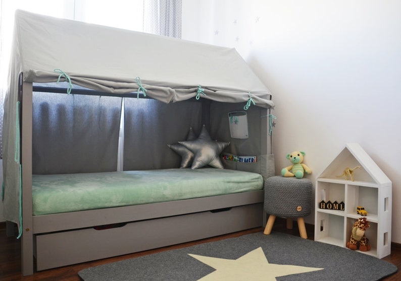 K&ouml;nnte beinhalten: Ein graues Holzbettgestell mit einem wei&szlig;en Baldachin und einer hellgr&uuml;nen Matratze. Das Bett hat eine Schublade darunter und einen sternf&ouml;rmigen Teppich davor. Es gibt einen grauen Strickhocker mit einem Teddyb&auml;r darauf und ein wei&szlig;es Holzregal mit dem Wort "HOME" darauf.