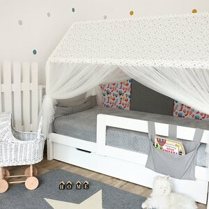 Puede incluir: Estructura de cama de madera blanca con dosel blanco y ropa de cama con estampado gris y blanco. La cama tiene un caj&oacute;n de almacenamiento y un bolsillo de tela gris en el lateral. La cama est&aacute; en una habitaci&oacute;n con una valla de madera blanca y un cochecito de mu&ntilde;ecas de mimbre blanco.