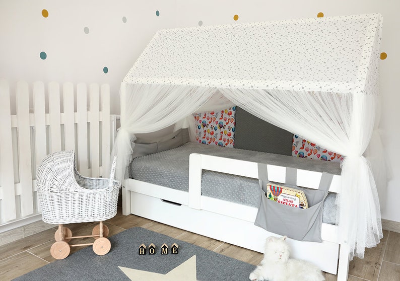 Puede incluir: Estructura de cama de madera blanca con dosel blanco y ropa de cama con estampado gris y blanco. La cama tiene un caj&oacute;n de almacenamiento y un bolsillo de tela gris en el lateral. La cama est&aacute; en una habitaci&oacute;n con una valla de madera blanca y un cochecito de mu&ntilde;ecas de mimbre blanco.