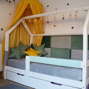 Könnte beinhalten: Ein weißes Holzbettgestell mit einem gelben Baldachin und einem Kopfteil mit grünem und grauem Muster. Das Bett hat eine hellblaue Matratze und eine graue und weiße gemusterte Decke. Es gibt mehrere sternförmige Kissen auf dem Bett. Das Bett hat zwei Schubladen darunter.