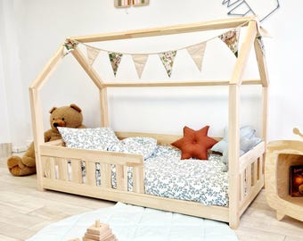 Könnte beinhalten: Ein hölzerner Haus-förmiger Bettrahmen mit einem weißen und grünen floralen Bettwäsche-Set. Das Bett hat ein Sicherheitsgeländer auf einer Seite und ein sternförmiges Kissen. Eine Schnur mit Stofffahnen hängt über dem Bett.