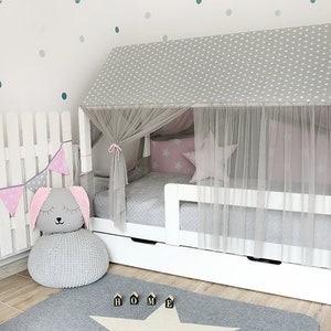 Puede incluir: Una cama de madera blanca con un dosel estampado en gris y blanco y una cortina de tul blanca. La cama tiene un colch&oacute;n gris y un caj&oacute;n blanco debajo. Hay un puf de punto gris con una cabeza de conejo gris en la parte superior, una pancarta de banderas rosa y gris y una alfombra gris con una estrella blanca.