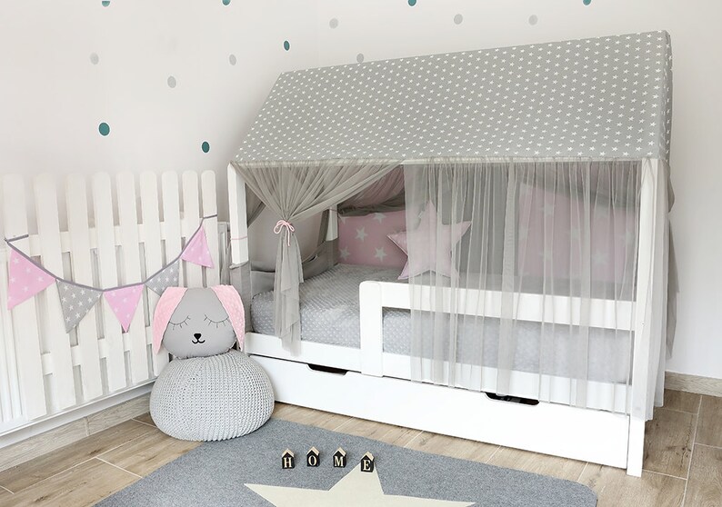 Puede incluir: Una cama de madera blanca con un dosel estampado en gris y blanco y una cortina de tul blanca. La cama tiene un colch&oacute;n gris y un caj&oacute;n blanco debajo. Hay un puf de punto gris con una cabeza de conejo gris en la parte superior, una pancarta de banderas rosa y gris y una alfombra gris con una estrella blanca.