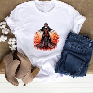 Könnte beinhalten: Weißes T-Shirt mit einer Aquarell-Illustration einer Hexe in einem schwarzen Kleid und einem roten Umhang, die in einem Feld mit roten Blumen steht.