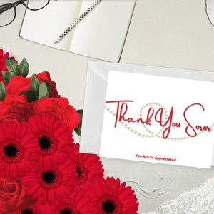 May include: A white greeting card with the words "Thank You Soror" in red script, with a pearl design. The card is next to a white envelope and a bouquet of red flowers, including roses and gerbera daisies.