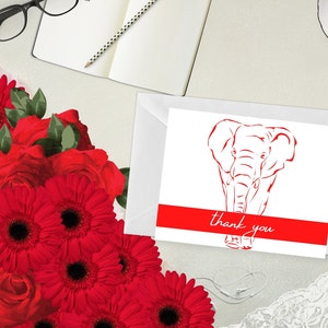May include: A white greeting card featuring a red elephant illustration and the words "thank you". The card is in a white envelope. Red flowers and a notebook with a pencil are in the background.