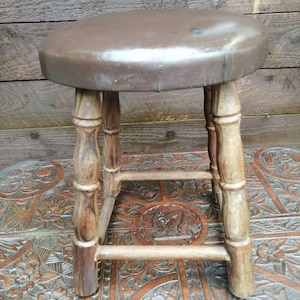 Puede incluir: Un taburete de madera marrón con un asiento redondo de cuero marrón. El taburete tiene cuatro patas con detalles torneados y un diseño sencillo y rústico.