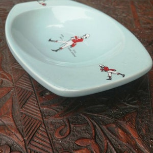 May include: A light blue ceramic ashtray with a white rim. The ashtray features a black and white illustration of a man in a red coat and white pants holding a riding crop. The text "Johnnie Walker" is printed on the ashtray.