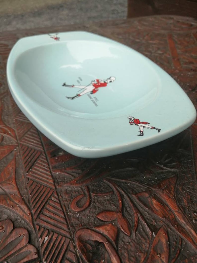 May include: A light blue ceramic ashtray with a white rim. The ashtray features a black and white illustration of a man in a red coat and white pants holding a riding crop. The text "Johnnie Walker" is printed on the ashtray.