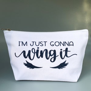 May include: White canvas makeup bag with a zipper closure. The bag has black glitter lettering that says "I'm just gonna wing it" with a pair of eyelashes on either side of the text.