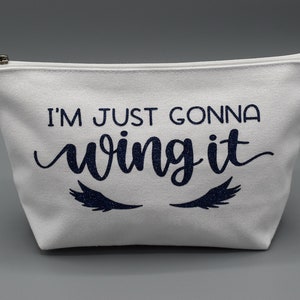 May include: White canvas zippered pouch with the text "I'm just gonna wing it" in navy blue with glitter eyelashes on either side.