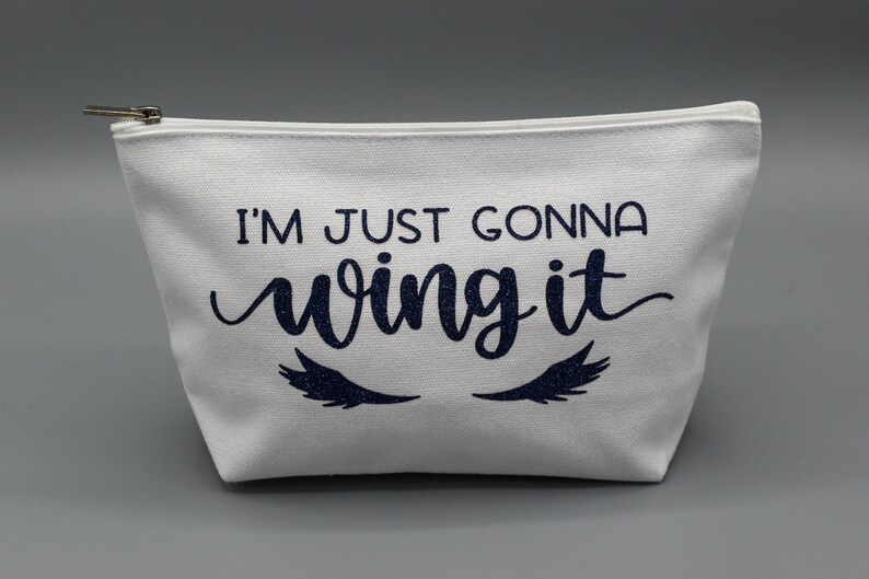 May include: White canvas zippered pouch with the text "I'm just gonna wing it" in navy blue with glitter eyelashes on either side.