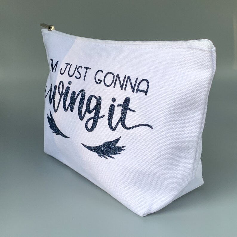 May include: White canvas makeup bag with a zipper closure. The bag has a black glitter design that says "I'm just gonna wing it" with two eyelashes on either side of the text.