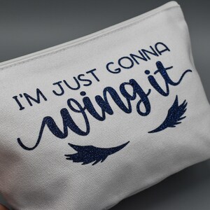 May include: White canvas zippered pouch with the text "I'm just gonna wing it" in blue glitter lettering. Two blue glitter eyelashes are below the text.