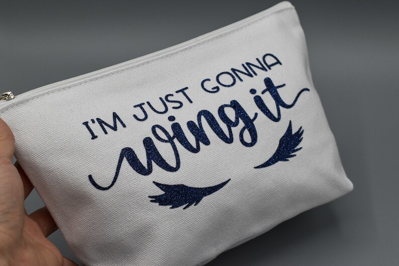 May include: White canvas zippered pouch with the text "I'm just gonna wing it" in blue glitter lettering. Two blue glitter eyelashes are below the text.